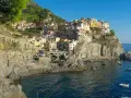 Le Cinque Terre, con le loro case colorate aggrappate alle scogliere, si affacciano sul mare blu. Gente prende il sole sulle rocce.