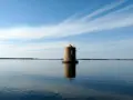Mulino solitario nella laguna di Orbetello, riflesso sull'acqua calma sotto un cielo azzurro con nuvole sottili.