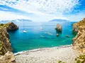 Rocky coastline with a pebble beach, turquoise water, sailboats, and distant islands under a blue, cloudy sky.