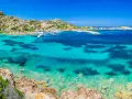 Acque cristalline e rocce granitiche sull'isola della Maddalena. Un porto con yacht e un hotel si affacciano sul mare turchese.
