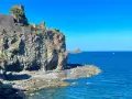 Castello Normanno a Aci Trezza, un luogo da vedere. Roccioso promontorio con antiche mura, mare blu e faraglioni all'orizzonte.