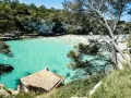 Una baia con acque turchesi cristalline, sabbia bianca e vegetazione lussureggiante. Un piccolo edificio con tetto di tegole si affaccia sul mare. Tra le più belle spiagge di Palma di Maiorca.