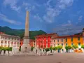 Piazza colorata con obelisco e leoni, incorniciata da edifici vivaci e montagne verdi, nella provincia di Massa e Carrara.