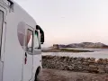 Un camper bianco parcheggiato su un lastricato di pietra, con una vista mozzafiato sul mare e una torre antica in lontananza. Un sogno di **Sardegna in camper**.