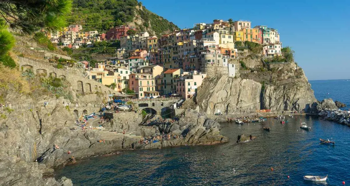 Le Cinque Terre, con le loro case colorate aggrappate alle scogliere, si affacciano sul mare blu. Gente prende il sole sulle rocce.