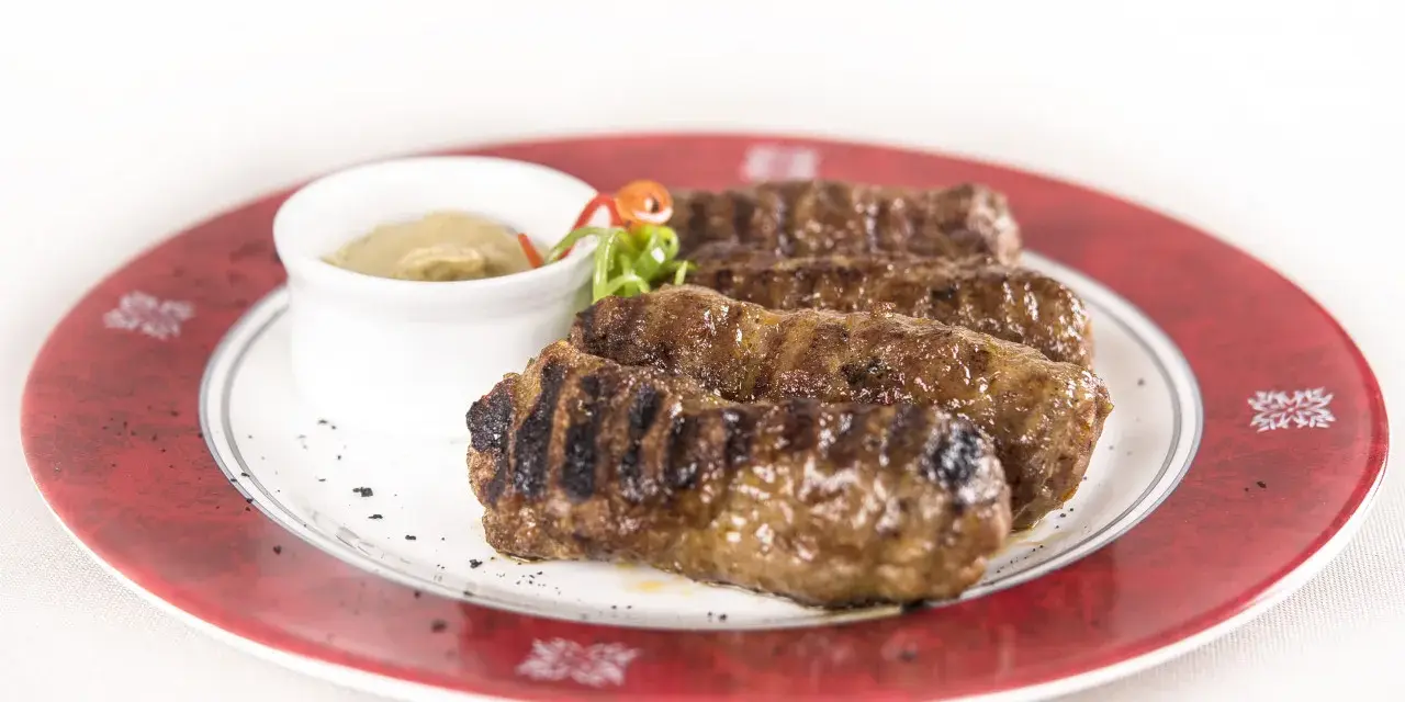 Mici grigliati su un piatto rosso, un classico per sapere cosa mangiare a Bucarest. Serviti con salsa.