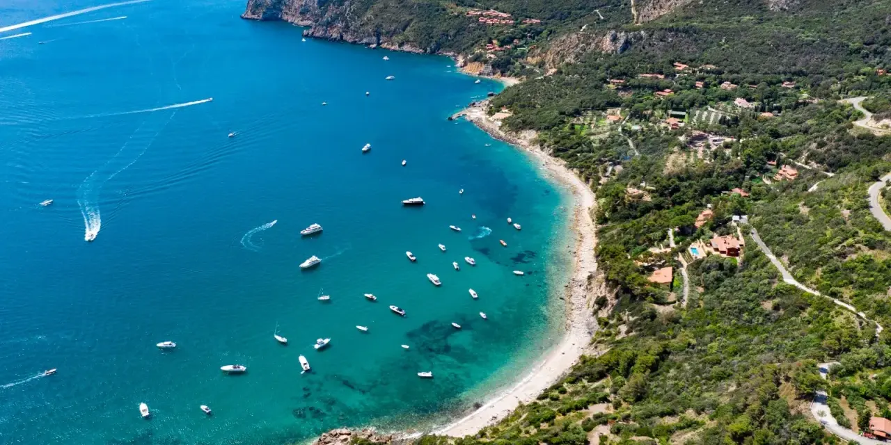 Baia con acque cristalline e numerose barche, incastonata nella costa verdeggiante dell'Argentario, dove si trova un paradiso naturale.