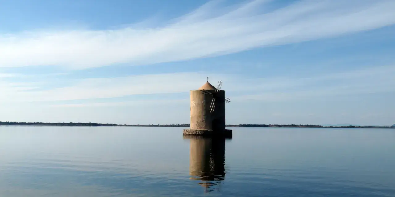 Mulino solitario nella laguna di Orbetello, riflesso sull'acqua calma sotto un cielo azzurro con nuvole sottili.