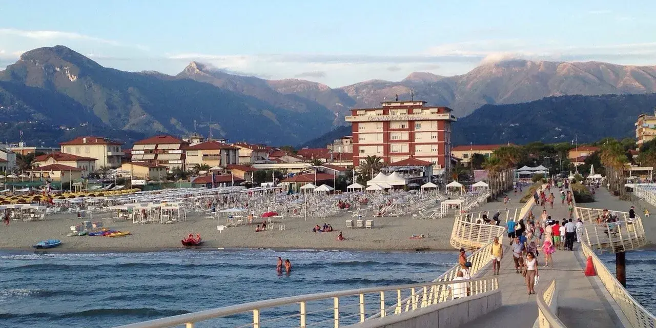 Passeggiata sul molo di Camaiore, dove si trova una spiaggia attrezzata con ombrelloni e montagne sullo sfondo.