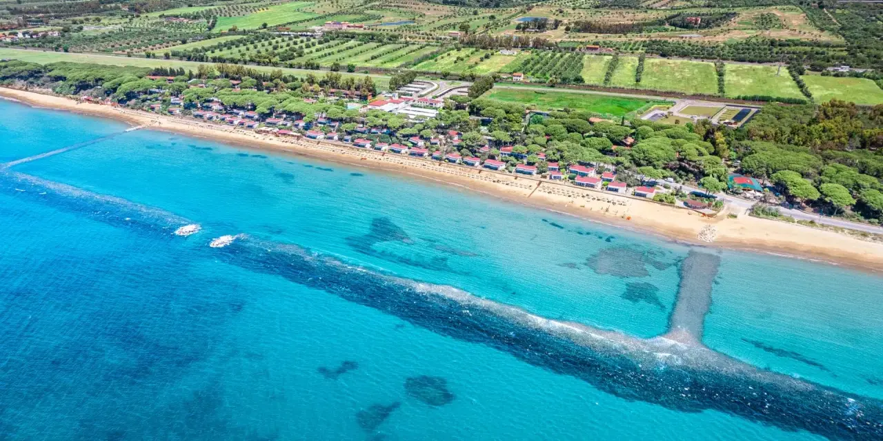Vista aerea di una spiaggia dorata e del mare cristallino a Castiglione della Pescaia, con pineta e campi verdi sullo sfondo.