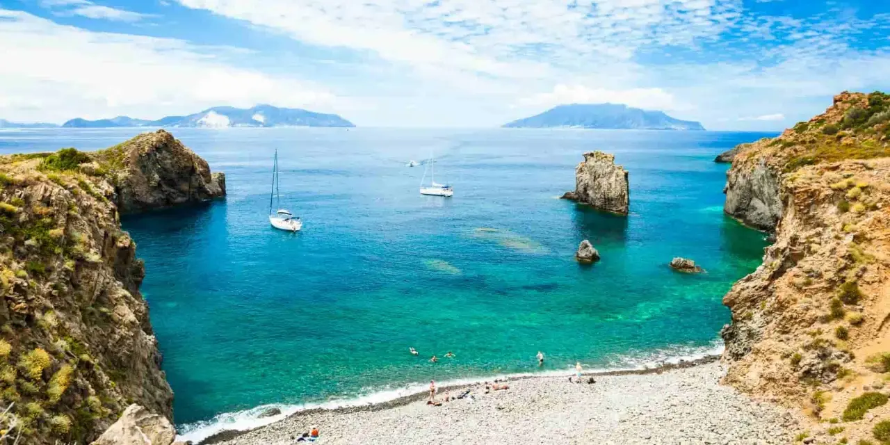 Rocky coastline with a pebble beach, turquoise water, sailboats, and distant islands under a blue, cloudy sky.
