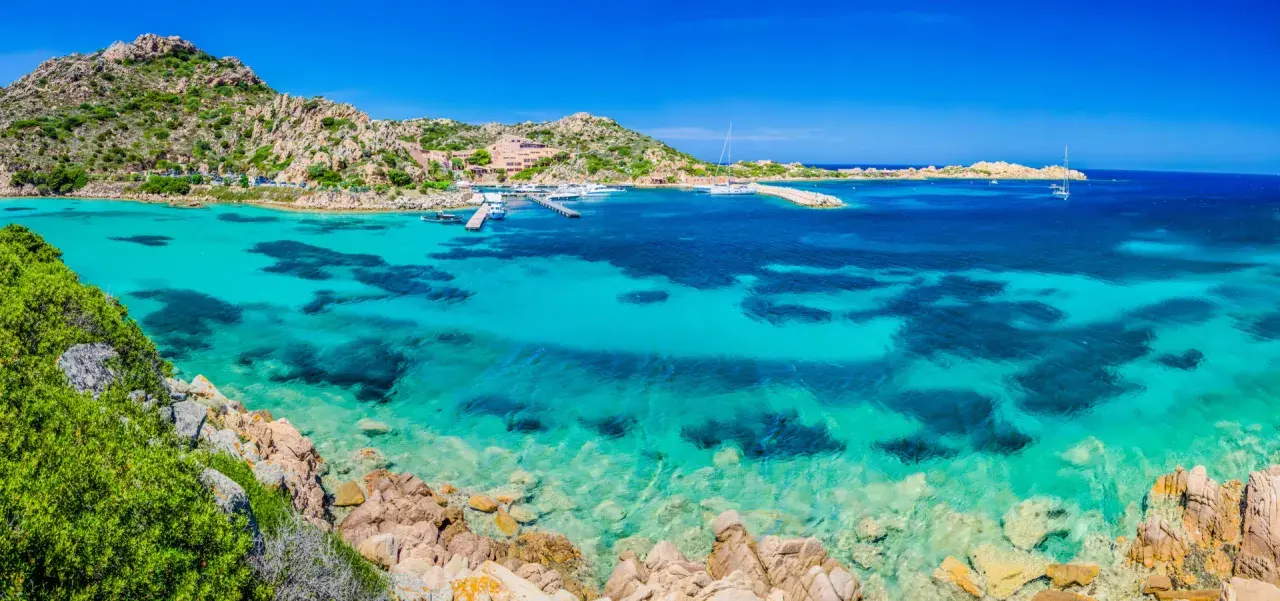 Acque cristalline e rocce granitiche sull'isola della Maddalena. Un porto con yacht e un hotel si affacciano sul mare turchese.