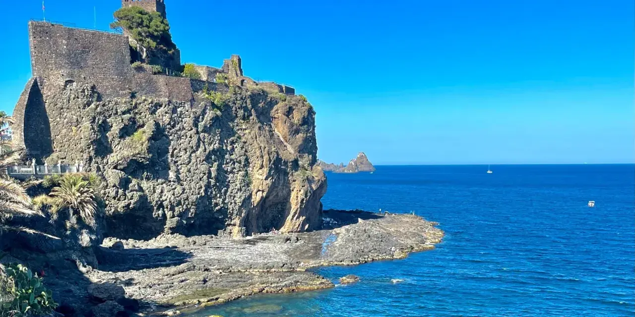 Castello Normanno a Aci Trezza, un luogo da vedere. Roccioso promontorio con antiche mura, mare blu e faraglioni all'orizzonte.