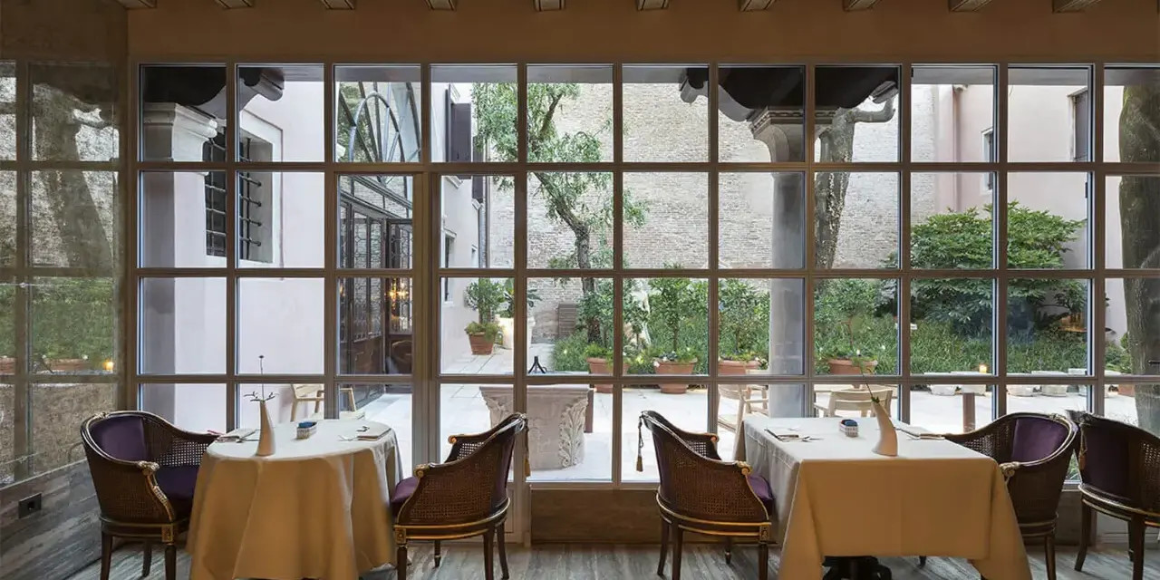 Tavoli apparecchiati in un elegante ristorante stellato Veneto, con vista su un cortile interno con alberi e piante in vaso.