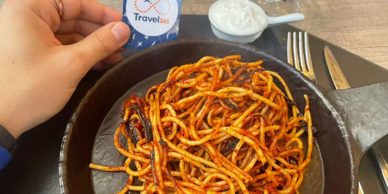 A hand holds a Travel365 tag over a plate of spaghetti with red sauce.