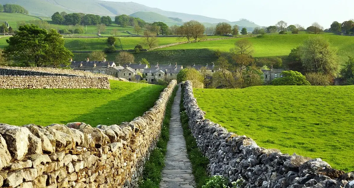 Sentiero tra muretti a secco in Yorkshire, con case di pietra e dolci colline verdi.