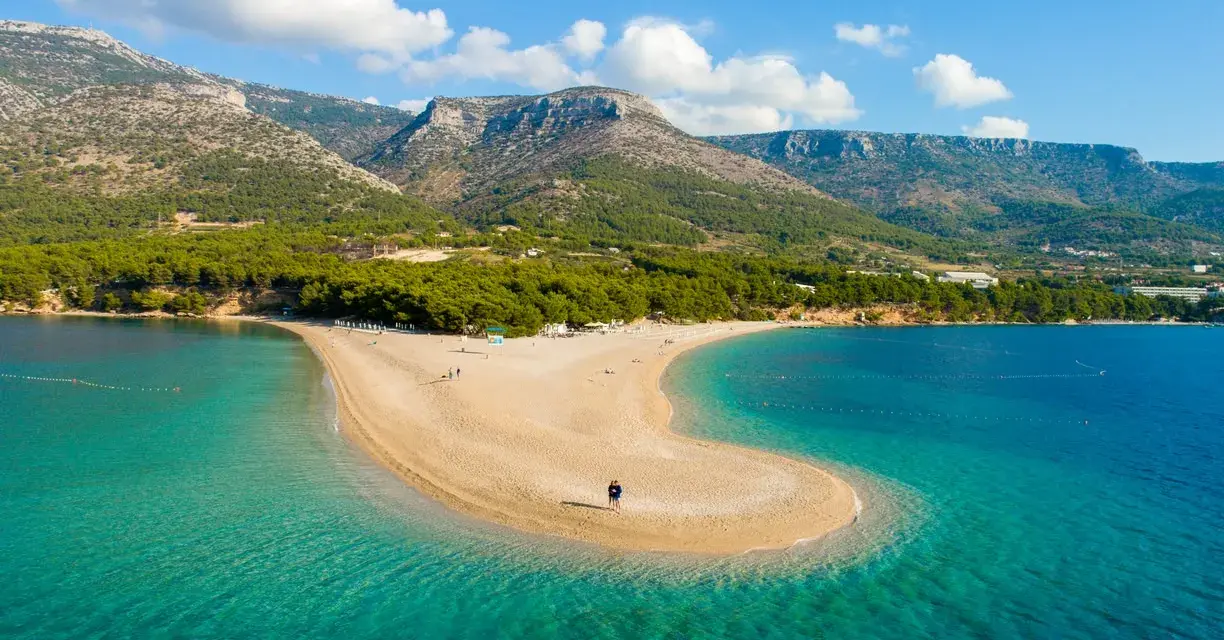 La Croazia offre il mare più bello: una spiaggia dorata a forma di lingua si protende in acque cristalline, con montagne verdi sullo sfondo.