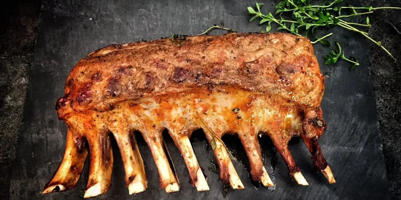 Carré d'agnello arrosto, uno dei 5 piatti tipici della Sardegna, guarnito con erbe aromatiche su una pietra scura.