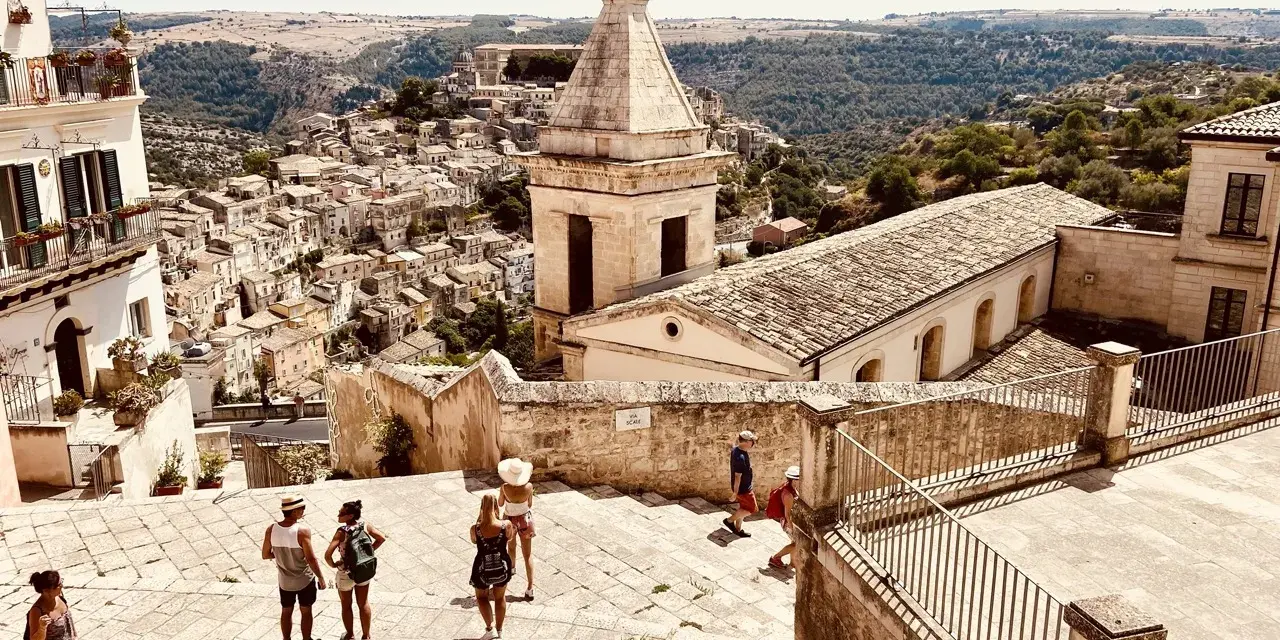 Panoramica di una città barocca in Sicilia orientale, cosa vedere: un borgo arroccato con chiese e case in pietra, turisti che esplorano le scalinate.