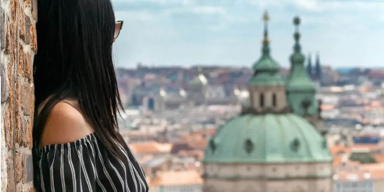 Vista di una donna con capelli scuri appoggiata a un muro di mattoni, con lo sguardo rivolto verso un panorama urbano di **città europee da visitare in 3 giorni**.