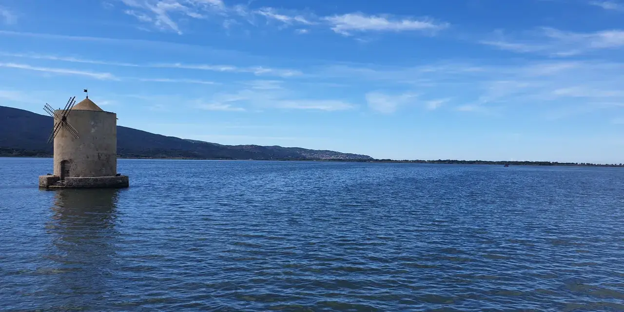 Mulino a Orbetello, un'icona da vedere. Paesaggio lagunare con cielo azzurro e colline sullo sfondo.