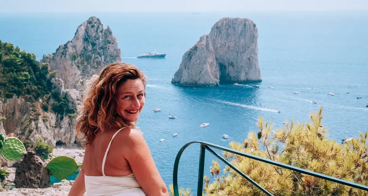 Donna sorridente con vista sui Faraglioni, un'icona dell'isola di Capri. Il mare azzurro e le barche completano il paesaggio.