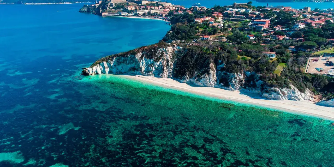 Vista aerea di una spiaggia bianca e mare cristallino, con un borgo arroccato su una collina. Un angolo di paradiso tra le isole arcipelago toscano.