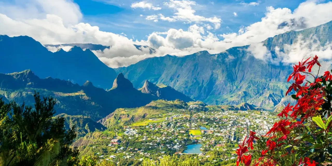 Vista panoramica di un villaggio verdeggiante sull'isola di Reunion, incastonato tra imponenti montagne blu e fiori rossi vibranti.