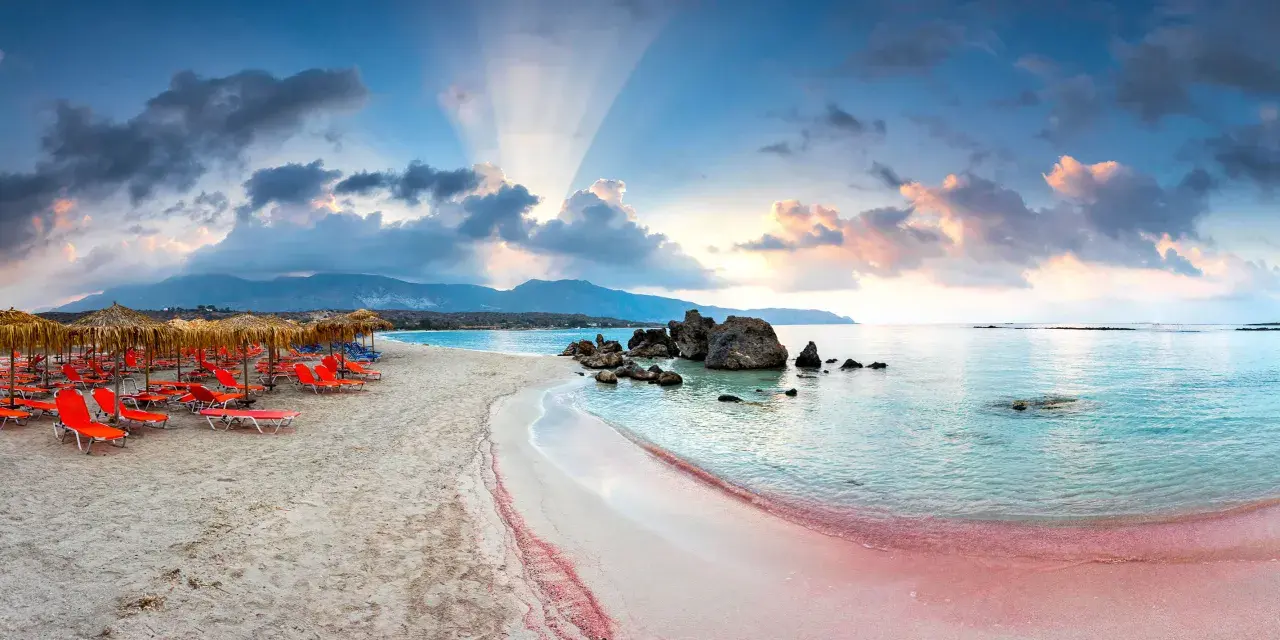 Spiaggia rosa con lettini arancioni e ombrelloni di paglia, mare turchese e montagne sullo sfondo. Un luogo da sogno tra i 10 mari più belli del mondo.