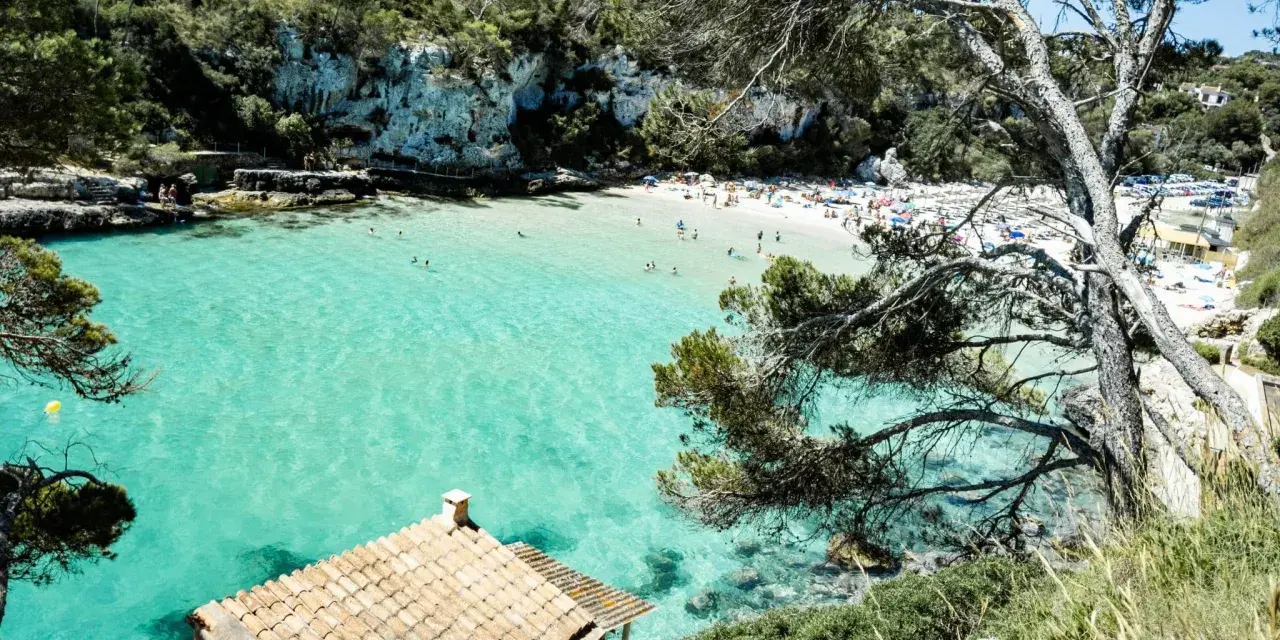 Una baia con acque turchesi cristalline, sabbia bianca e vegetazione lussureggiante. Un piccolo edificio con tetto di tegole si affaccia sul mare. Tra le più belle spiagge di Palma di Maiorca.