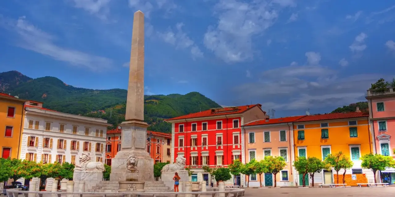 Piazza colorata con obelisco e leoni, incorniciata da edifici vivaci e montagne verdi, nella provincia di Massa e Carrara.