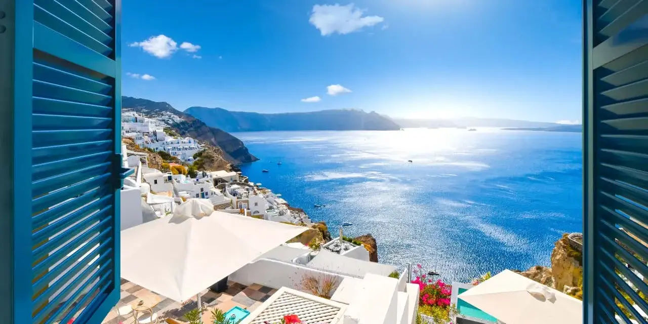 Vista mozzafiato su Santorini, perfetta per sapere dove andare in vacanza a giugno. Case bianche a picco sul mare blu intenso, fiori colorati e sole splendente.