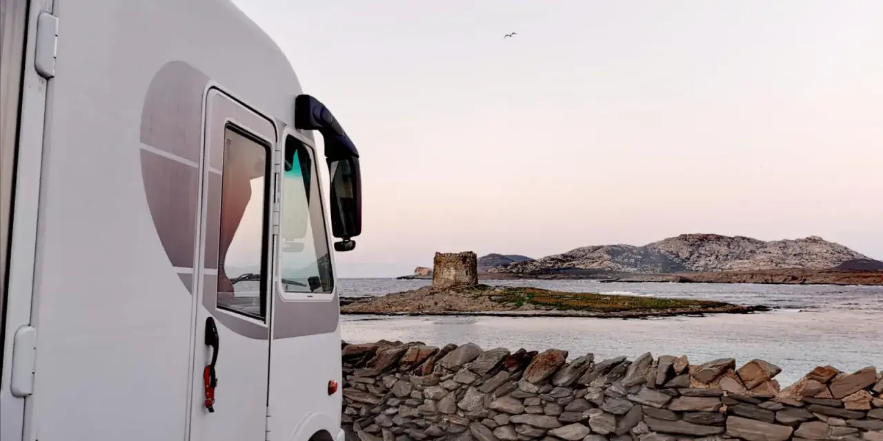 Un camper bianco parcheggiato su un lastricato di pietra, con una vista mozzafiato sul mare e una torre antica in lontananza. Un sogno di **Sardegna in camper**.