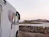 Un camper bianco parcheggiato su un lastricato di pietra, con una vista mozzafiato sul mare e una torre antica in lontananza. Un sogno di **Sardegna in camper**.