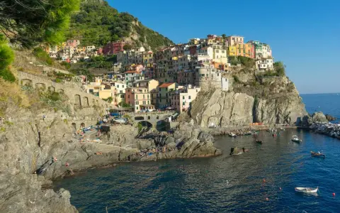 Le Cinque Terre, con le loro case colorate aggrappate alle scogliere, si affacciano sul mare blu. Gente prende il sole sulle rocce.
