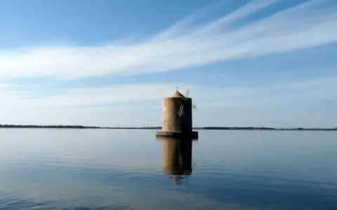 Mulino solitario nella laguna di Orbetello, riflesso sull'acqua calma sotto un cielo azzurro con nuvole sottili.
