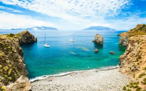 Rocky coastline with a pebble beach, turquoise water, sailboats, and distant islands under a blue, cloudy sky.