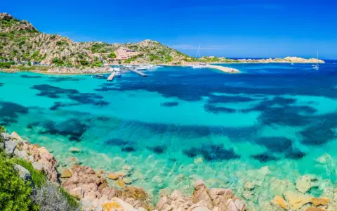 Acque cristalline e rocce granitiche sull'isola della Maddalena. Un porto con yacht e un hotel si affacciano sul mare turchese.