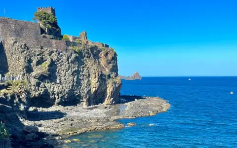Castello Normanno a Aci Trezza, un luogo da vedere. Roccioso promontorio con antiche mura, mare blu e faraglioni all'orizzonte.
