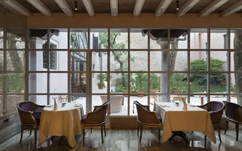 Tavoli apparecchiati in un elegante ristorante stellato Veneto, con vista su un cortile interno con alberi e piante in vaso.