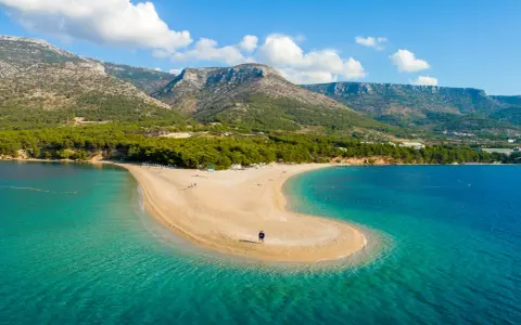 La Croazia offre il mare più bello: una spiaggia dorata a forma di lingua si protende in acque cristalline, con montagne verdi sullo sfondo.