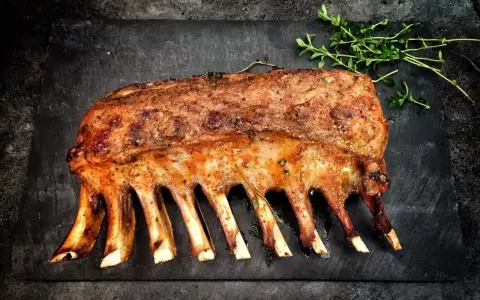 Carré d'agnello arrosto, uno dei 5 piatti tipici della Sardegna, guarnito con erbe aromatiche su una pietra scura.