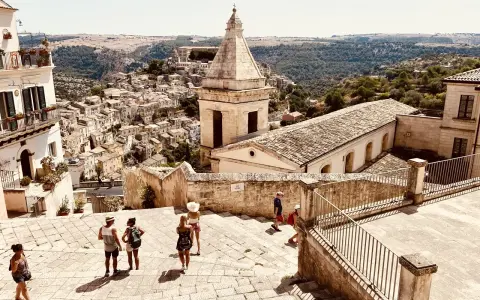 Panoramica di una città barocca in Sicilia orientale, cosa vedere: un borgo arroccato con chiese e case in pietra, turisti che esplorano le scalinate.