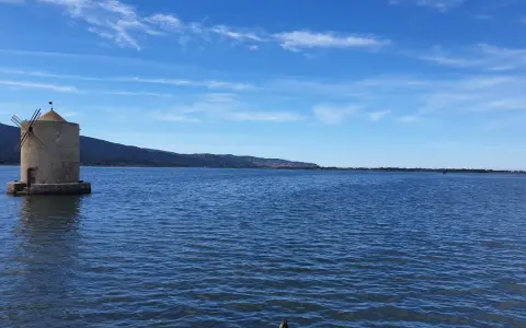 Mulino a Orbetello, un'icona da vedere. Paesaggio lagunare con cielo azzurro e colline sullo sfondo.