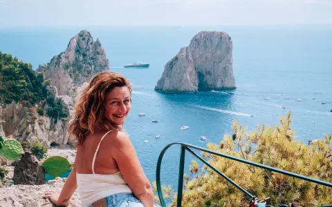 Donna sorridente con vista sui Faraglioni, un'icona dell'isola di Capri. Il mare azzurro e le barche completano il paesaggio.