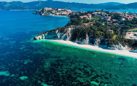 Vista aerea di una spiaggia bianca e mare cristallino, con un borgo arroccato su una collina. Un angolo di paradiso tra le isole arcipelago toscano.