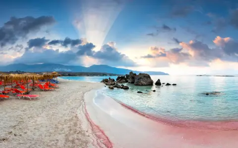 Spiaggia rosa con lettini arancioni e ombrelloni di paglia, mare turchese e montagne sullo sfondo. Un luogo da sogno tra i 10 mari più belli del mondo.