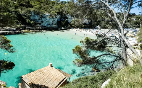Una baia con acque turchesi cristalline, sabbia bianca e vegetazione lussureggiante. Un piccolo edificio con tetto di tegole si affaccia sul mare. Tra le più belle spiagge di Palma di Maiorca.