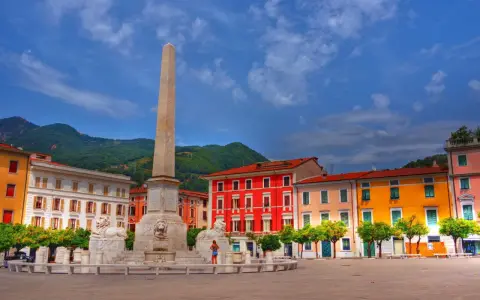 Piazza colorata con obelisco e leoni, incorniciata da edifici vivaci e montagne verdi, nella provincia di Massa e Carrara.