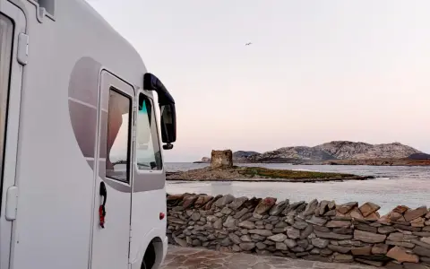 Un camper bianco parcheggiato su un lastricato di pietra, con una vista mozzafiato sul mare e una torre antica in lontananza. Un sogno di **Sardegna in camper**.